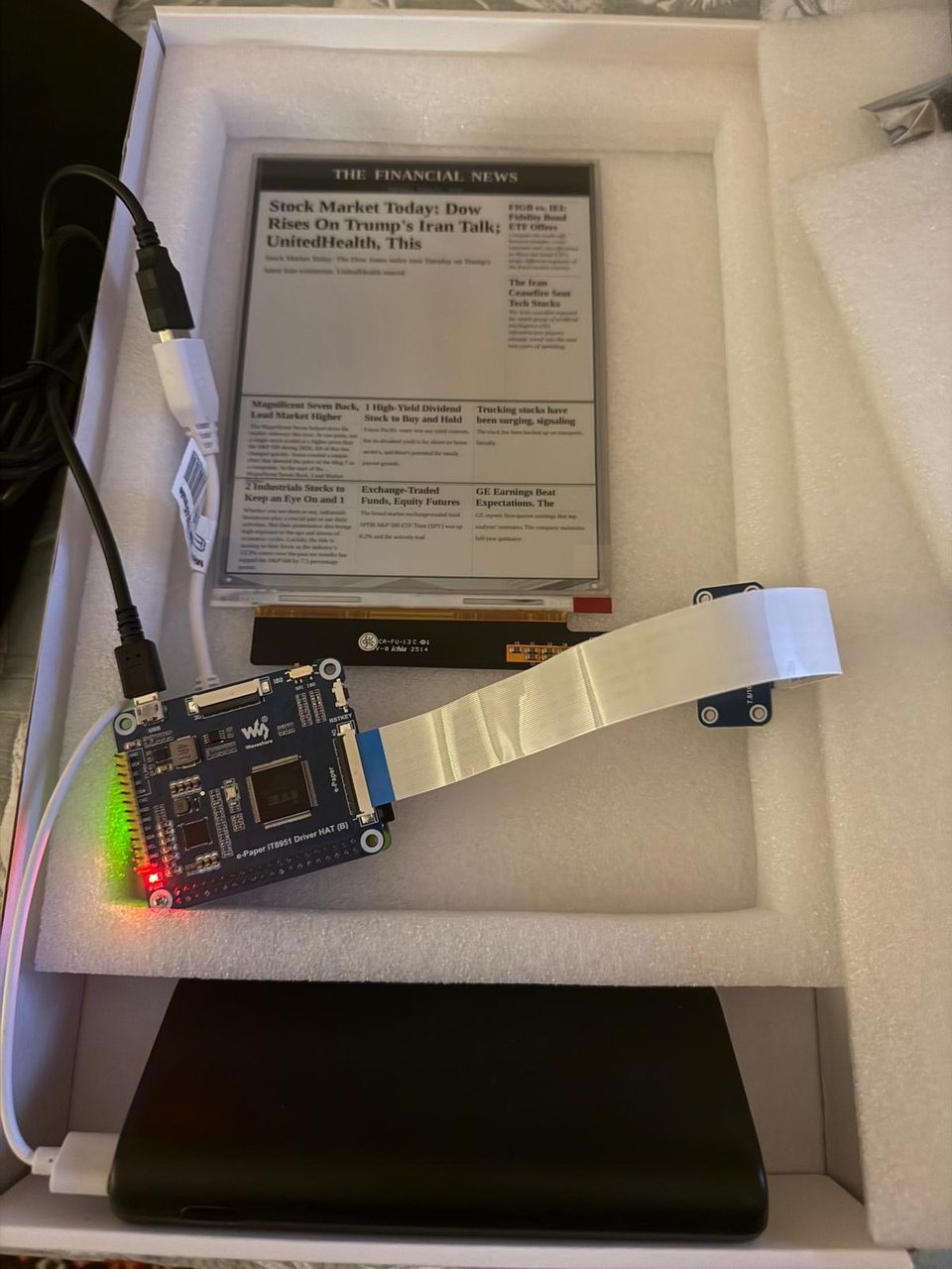 An IT8951 driver board with its status LEDs lit, a ribbon cable running to an e-ink panel showing a live front page, all sitting in a tray of packing foam next to a powerbank.