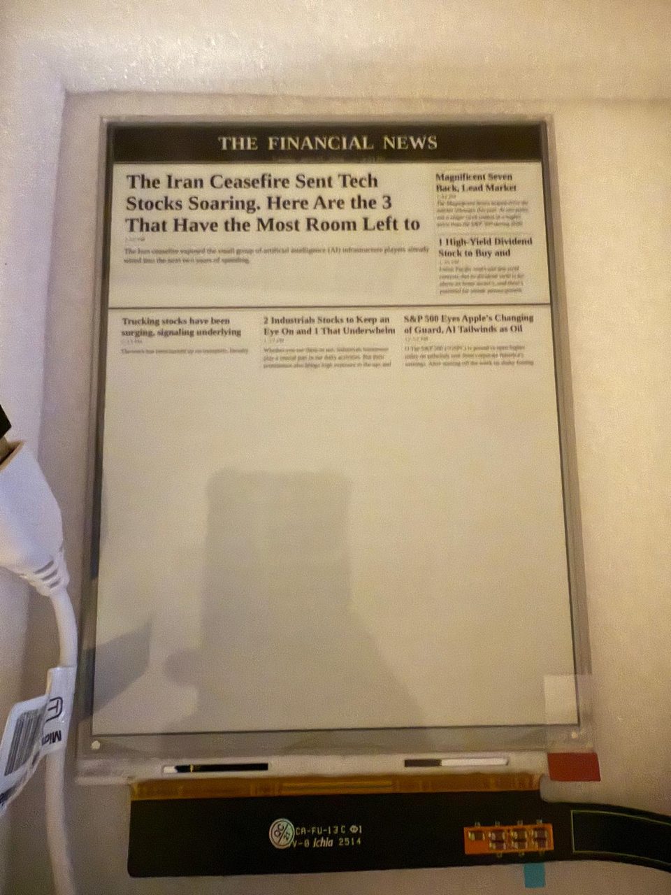 A bare 7.8-inch e-ink panel taped to a wall, showing a rendered front page headed THE FINANCIAL NEWS; a ribbon cable and driver board hang below.